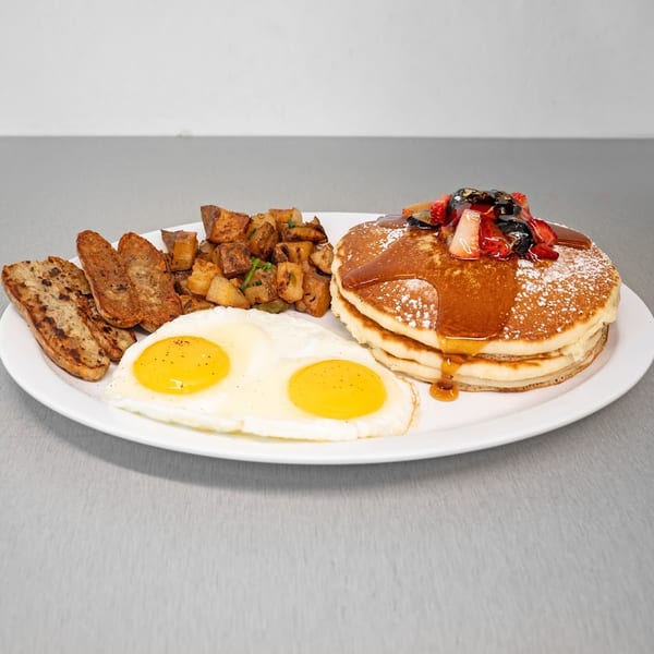 A breakfast plate with pancakes, eggs, sausage, and home fries on a white plate.
