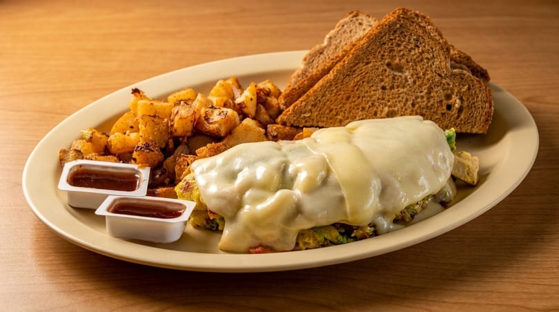 A breakfast platter with a cheese-covered omelet, home fries, two slices of toast, and two ketchup cups.