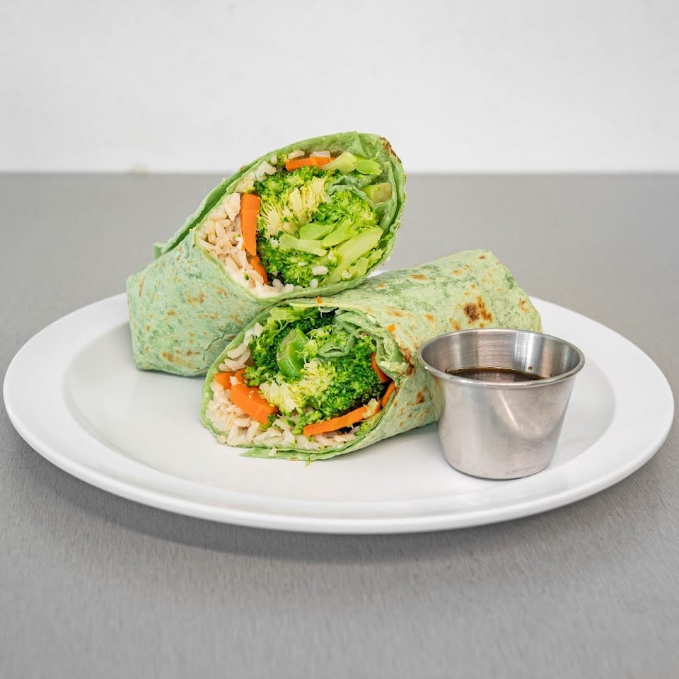 Two halves of a green tortilla wrap filled with broccoli, carrots, and rice, with a side of sauce.