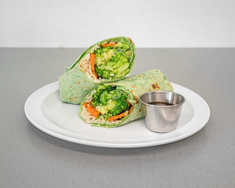 Two halves of a green tortilla wrap filled with broccoli, carrots, and rice, with a side of sauce.