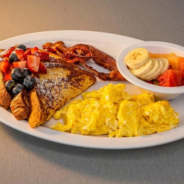 Breakfast plate with French toast, berries, bacon, scrambled eggs, and a fruit cup.