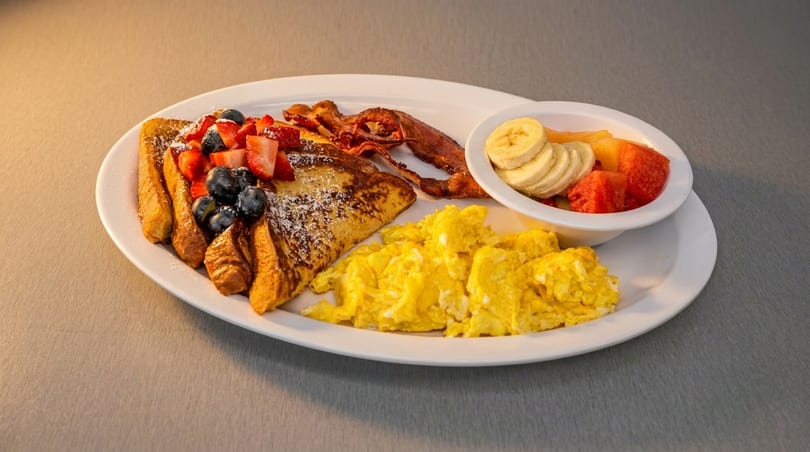Breakfast plate with French toast, berries, bacon, scrambled eggs, and a fruit cup.