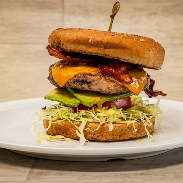 Tall bacon cheeseburger with avocado, tomato, and shredded lettuce on a white plate.
