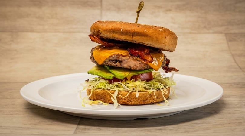 Tall bacon cheeseburger with avocado, tomato, and shredded lettuce on a white plate.