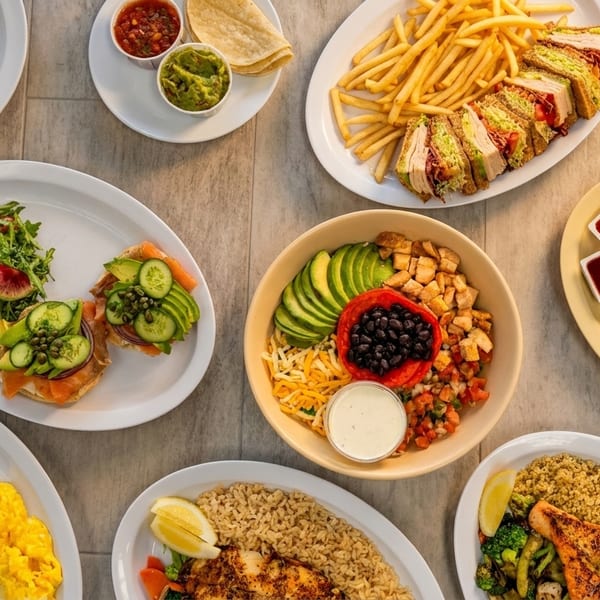 An overhead shot of various dishes, including wraps, sandwiches, salads, and entrees, on a light-colored table.