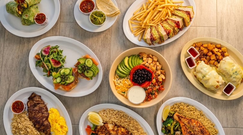 An overhead shot of various dishes, including wraps, sandwiches, salads, and entrees, on a light-colored table.