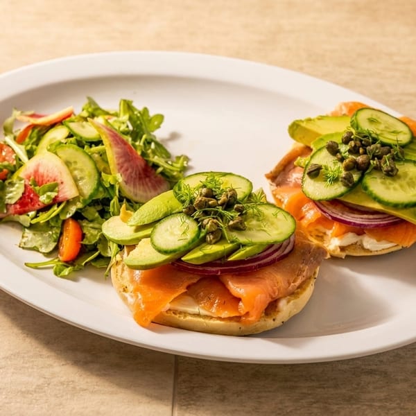 Two bagels with lox, avocado, cucumber, red onion, and capers, served with a side salad.