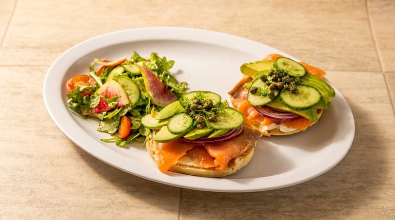 Two bagels with lox, avocado, cucumber, red onion, and capers, served with a side salad.